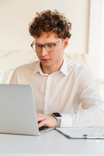 Confident attractive young businessman wearing white shirt working on laptop computer while sitting at the table at home